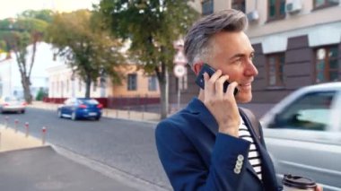 Beyaz tenli yakışıklı orta yaşlı iş adamı sokakta yürüyor, kahve içiyor ve cep telefonuyla konuşuyor. Dolaşan ve cep telefonuyla konuşan bir adam. Sıcak bir şeyler içiyorum. Meşgul erkek telefonda arıyor..