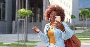Afro-Amerikan güzel, mutlu, gülümseyen kadın dışarıda cep telefonundan web kamerasıyla konuşuyor. Neşeli stil sahibi kıvırcık kadın cep telefonuyla konuşuyor ve gülümsüyor. Görüntülü konuşma. Video Hattı.