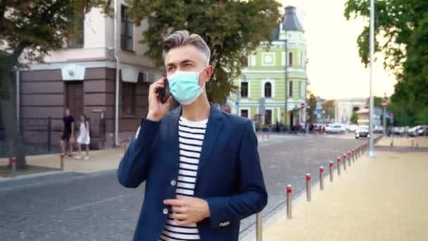 Hombre de negocios guapo caucásico con máscara médica caminando por la calle de la ciudad y hablando por teléfono. Un hombre guapo paseando al aire libre y hablando por celular. Tengo una llamada de negocios. pandemia de Covid-19. — Vídeos de Stock