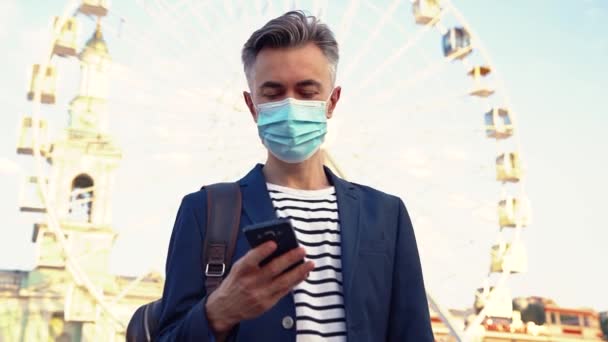 在城里戴着医疗面具的白种人英俊男子，并在手机上发短信。漂亮的商人站在外面的摩天轮上，用智能手机敲敲或滚动。Coronavirus时间. — 图库视频影像