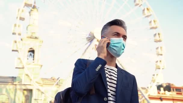 Hombre de negocios guapo caucásico en protección respiratoria en la calle de la ciudad hablando por teléfono móvil. Hombre guapo con máscara parado en la noria y hablando en el teléfono inteligente. Conversación telefónica. — Vídeo de stock