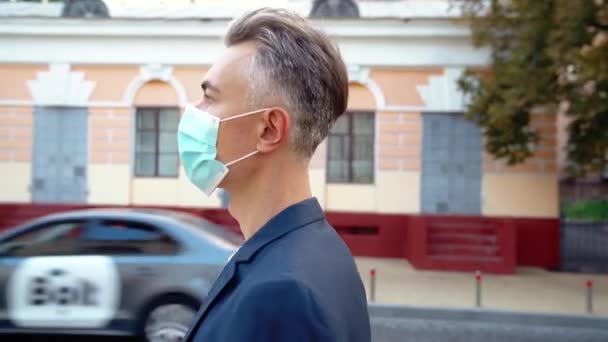 Side view on Caucasian middle-aged handsome man in medical mask walking the street in town, turning face and looking at camera. Businessman in respiratory protection stepping outside. Close up. — Stock Video