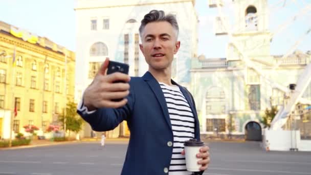 Hombre de negocios caucásico guapo de mediana edad de pie en la calle de la ciudad, bebiendo café y tomando una foto selfie en el teléfono móvil. Hombre alegre haciendo fotos en el teléfono inteligente. Bebiendo bebida caliente. Exterior. — Vídeos de Stock