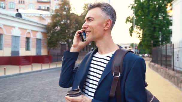 Кавказький гарний бізнесмен стоїть на вулиці, п'є каву і розмовляє з посмішкою на мобільному телефоні. Чоловік говорить по мобільному на вулиці. Гарячий напій. Зайнятий чоловік розмовляє по телефону.. — стокове відео