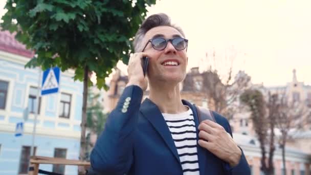 Kaukázusi vidám üzletember napszemüvegben áll az utcán és mosolyog a mobilján. Mosolygó férfi beszél mobilon Elfoglalt boldog férfi szemüvegben csevegő telefonon és mosolygós — Stock videók