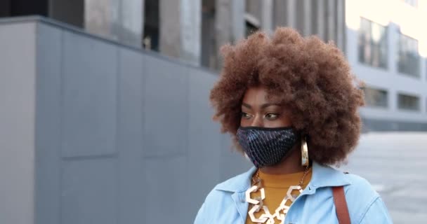 Africano americano jovem elegante mulher bonita com peruca cabelo encaracolado e na máscara andando na rua, batendo e rolando no smartphone. Linda mulher ao ar livre na cidade mensagem de texto no telefone móvel. — Vídeo de Stock