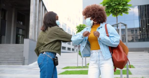 Genç, Afro-Amerikalı, tıbbi maskeli en iyi arkadaşlar sokakta buluşup dirseklerle selamlaşıyorlar. Mutlu kızlar neşe içinde konuşup sohbet ediyorlar. Koronavirüs salgını sırasında öğrenciler. — Stok video