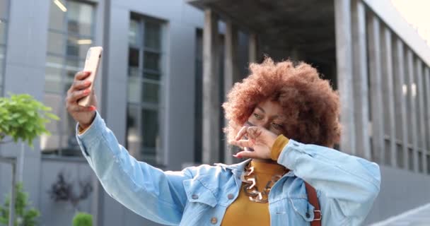 Afro-américaine jeune jolie femme hipster élégant avec perruque bouclée et dans le masque posant à la caméra smartphone et de prendre des photos selfie dans la rue urbaine. Belle femme en plein air en ville faisant des photos. — Video