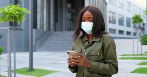 Afro-Américain jeune jolie femme élégante dans le masque médical debout dans la rue urbaine et en utilisant un smartphone. Belle femme en plein air en ville textos message sur téléphone mobile. Pandémie de coronavirus. — Video