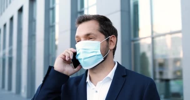 Homem alegre bonito jovem caucasiano na máscara médica falando no telefone celular e sorrindo do lado de fora no centro de negócios. Homem de negócios feliz na proteção respiratória falando no celular. Pandemia. — Vídeo de Stock