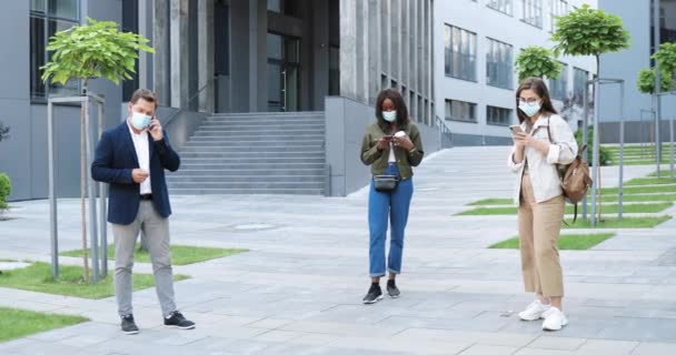 Karışık ırk erkek ve kadın tıbbi maskeli açık havada duran ve alet kullanan. İş adamı cep telefonuyla konuşuyor, kadınların mesajlaşması, dinlemesi ve akıllı telefonları kaydırması. Salgın sosyal mesafesi. — Stok video