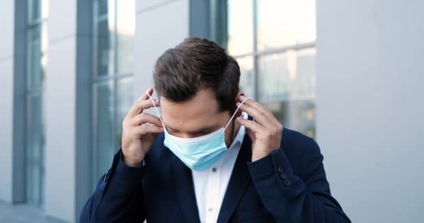 Portrait of young Caucasian handsome man taking on medical mask and looking at camera outdoor. Attractive stylish businessman wearing respiratory protection at street at business center in quarantine — Stock Video