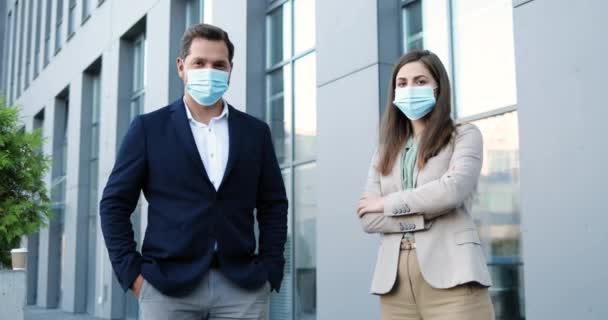 Portret van een blanke jonge man en vrouw in zakelijke stijl en in medische maskers die op sociale afstand staan en naar de camera kijken. Zakenman en zakenvrouw buiten tijdens pandemie. — Stockvideo