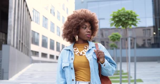 Portrait tourné de jeune afro-américaine jolie femme hipster en jean veste debout dans la rue de la ville, tournant le visage et souriant joyeusement à la caméra. Belle bouclé heureux gai femme en plein air. — Video