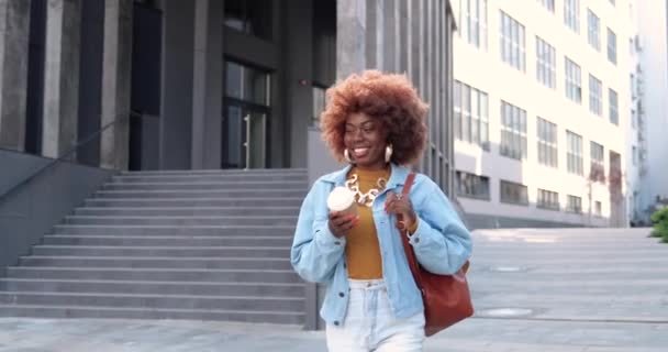 Jonge stijlvolle Afro-Amerikaanse mooie krullende vrouw met tas lopen in de stad straat, met een kopje koffie-to-go en glimlachen vrolijk. Vrij gelukkig vrouwtje wandelen buiten met koffie en lachen. — Stockvideo