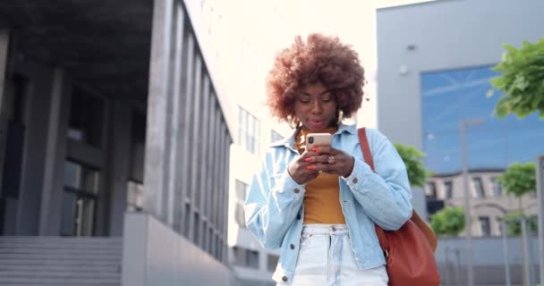 Afro-américaine jeune femme bouclée élégant tapotant ou défilant sur smartphone et debout à la rue de la ville. Magnifique message texte féminin sur téléphone portable et bavardage. Extérieur. — Video