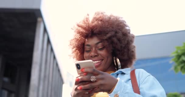 Primo piano di afro-americano gioioso giovane donna elegante toccando o scorrendo su smartphone e in piedi in strada della città. Bella felice messaggio di testo femminile sul telefono cellulare e sorridente. Fuori.. — Video Stock