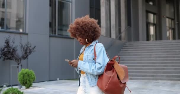 Afroamericana joven mujer rizada con estilo tocando o desplazándose en el teléfono inteligente y caminando en la calle de la ciudad. Hermoso mensaje de texto femenino en el teléfono móvil y pasear. Afuera. Mensajería. — Vídeo de stock