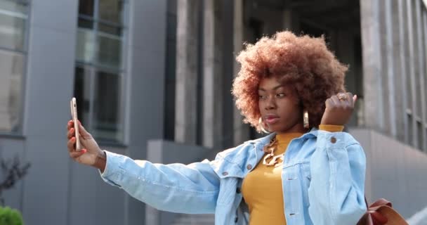 Belle jeune femme frisée afro-américaine posant sur un appareil photo smartphone tout en prenant des photos selfie dans la rue. Plutôt élégant cool femelle faisant selfies photos avec téléphone mobile. — Video