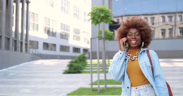 Nahaufnahme einer schönen lächelnden Afroamerikanerin, die mit ihrem Handy telefoniert und im Freien spazieren geht. Fröhliche Frauen, die am Handy sprechen und lachen. Nettes Telefonat. — Stockvideo