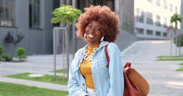 Porträt einer jungen hübschen Afroamerikanerin in Jeansjacke, die auf der Straße steht, lacht und freudig in die Kamera lächelt. Schöne lockige fröhliche Frau posiert draußen und zeigt Siegesgeste. — Stockvideo