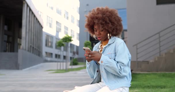 Mujer hermosa afroamericana tocando y desplazándose en el teléfono móvil y sentado en el banco fuera. Elegante mensajería femenina atractiva en el teléfono inteligente. Charlando por teléfono. — Vídeos de Stock