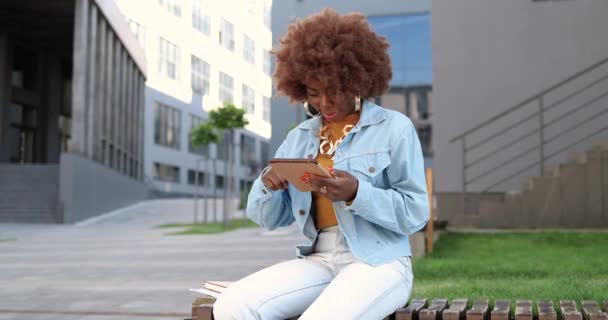 Afro-américaine belle femme tapotant et défilant sur tablette dispositif, souriant et assis sur le banc à l'extérieur. Élégant attrayant messagerie féminine et bavarder sur un ordinateur gadget en ville. — Video