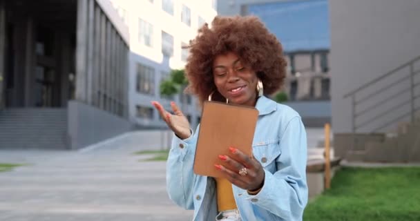 Jeune afro-américaine frisée belle femme assise sur un banc, parlant via webcam sur tablette et riant en plein air. Jolie vidéoconférence féminine sur gadget à la rue. Vidéochat. — Video