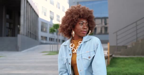 Portrait de jeune femme afro-américaine en veste de jeans debout dans la rue de la ville, tournant le visage et souriant à la caméra. Belle bouclé heureux gai femme en plein air. — Video