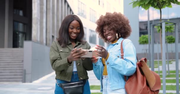 Les femmes afro-américaines parlent et regardent quelque chose sur leur téléphone portable dans la rue de la ville. Belles jeunes femmes avec une tasse de café à emporter en utilisant un smartphone. Des amis joyeux bavardant. Des commérages. — Video