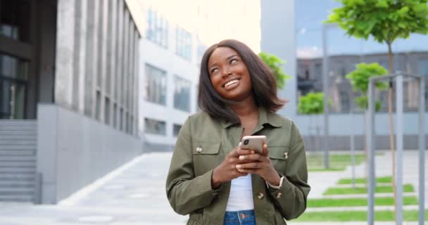 Афроамериканка радісна молода стильна жінка стукає або прокручується по смартфону і стоїть на міській вулиці. Чудова жінка пише смс на мобільному телефоні і посміхається. Зовні. — стокове відео