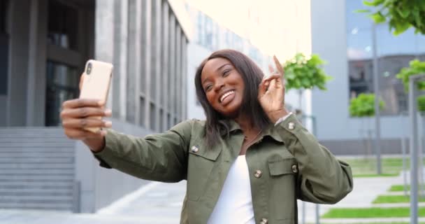 Belle jeune femme afro-américaine posant et souriant à la caméra du smartphone tout en prenant des photos selfie dans la rue. Jolie femme élégante gaie faisant selfies photos avec téléphone mobile. — Video