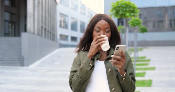 Afro-Américaine joyeuse jeune femme élégante regardant la vidéo sur smartphone et sirotant une boisson chaude, marchant le matin dans la rue. Belle femme heureuse utilisant téléphone portable et boire du café. Extérieur — Video