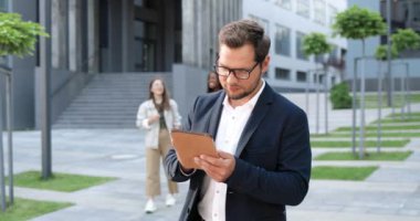 Beyaz tenli, genç, şık bir adam, sokaktaki tablet cihazını tıklatıyor ya da kaydırıyor. Yakışıklı erkek mesajlaşması ve alet bilgisayarı. Gözlüklü iş adamı dışarıda çalışıyor..