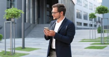 Kafkasyalı mutlu, genç, şık iş adamı akıllı telefonları dinliyor ya da kaydırıyor ve sokakta dikiliyor. Yakışıklı bir erkek telefonda mesaj atıyor ve gülümsüyor. Açık havada..
