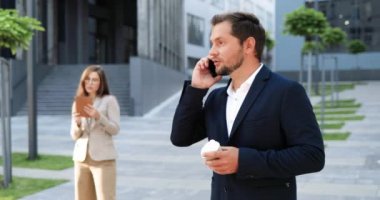 Beyaz, neşeli, genç, şık bir adam cep telefonuyla konuşuyor ve sabah sokakta sıcak bir şeyler yudumluyor. Yakışıklı mutlu erkek cep telefonuyla konuşuyor ve kahve içiyor. Dışarıda..