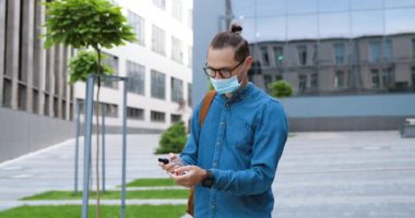 Gözlüklü ve tıbbi maskeli genç beyaz adam dışarıda duruyor ve cildine dezenfektan püskürtüyor. Gözlüklü yakışıklı erkek öğrenci dezenfektanla dezenfekte edip, ellerini temizliyor..