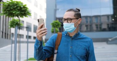 Gözlüklü ve tıbbi maskeli beyaz adam Steet 'teki cep telefonundan web kamerasıyla konuşuyor. Gözlüklü yakışıklı erkek dışarıda cep telefonuyla sohbet ediyor. Görüntülü konuşma. Video Hattı.