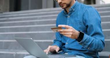 Gözlüklü ve kot pantolonlu beyaz bir adam kredi kartı kullanıyor ve internetten alışveriş yapıyor. Gözlüklü yakışıklı bir erkek bilgisayarda yazı yazıyor ve internetten alışveriş yapıyor. Sokakta alıcı.