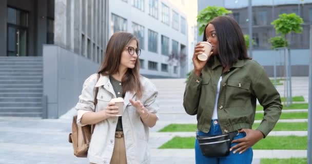 Μικτές φυλές νεαρές όμορφες γυναίκες, κολλητές που μιλάνε χαρούμενα και περπατούν με κούπες καφέ στο δρόμο της πόλης. Πολυεθνικές όμορφες, χαρούμενες φοιτήτριες βγαίνουν έξω με ποτά.. — Αρχείο Βίντεο