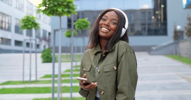 Joyeux jeune afro-américaine belle femme dans les écouteurs souriant joyeusement à la rue de la ville et écouter de la musique sur smartphone. Jolie femme heureuse appréciant la chanson sur le lecteur de téléphone mobile. — Video