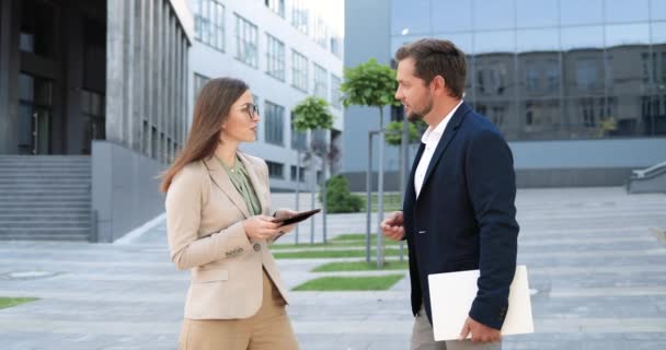 Caucásico hermosa mujer sosteniendo dispositivo de la tableta y hablando con el hombre guapo fuera. Hombre y feale socios de negocios reunión y teniendo discusión. Empresario y empresaria hablan en la calle. — Vídeos de Stock