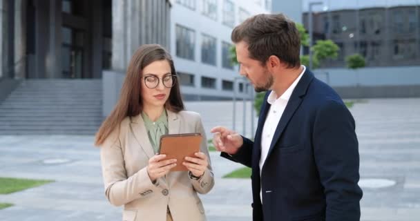 Beyaz erkek ve kadın meslektaşlar şehirde durup tablet cihazıyla ilgili bazı belgeler tartışıyorlar. İş ortakları konuşuyor ve dışarıda alet kullanıyorlar. Kadın ve erkek kasabada tartışıyorlar.. — Stok video