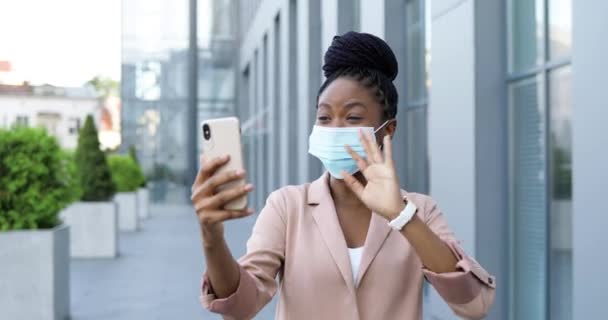 Ganska glad afroamerikansk ung kvinna i medicinsk mask som har videochatt på smartphone utomhus på business building. Glada vackra kvinnor som pratar och videochattar via webbkamera på mobiltelefon. — Stockvideo