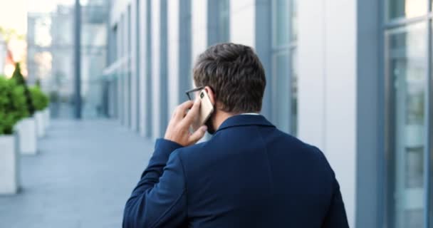 Arrière sur Caucasien jeune homme élégant parler sur téléphone portable et marcher à la ville. Beau mâle heureux se promenant en ville, parlant sur un téléphone portable. Un homme d'affaires répond au téléphone. Vue de derrière. À l'extérieur — Video