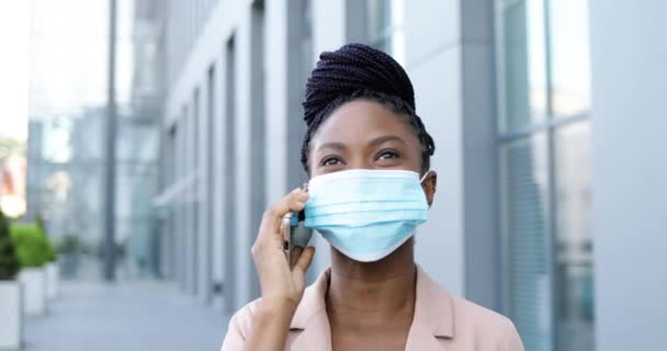 Afroamericana joven hermosa mujer alegre en máscara médica hablando por teléfono móvil y riendo afuera en el centro de negocios. Feliz empresaria en protección respiratoria hablando por celular. — Vídeos de Stock