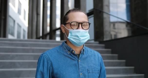 年轻英俊的男生戴着医疗面具和眼镜站在室外看着相机的画像。Coronavirus流行病的概念。戴眼镜站在街上的白人男子与保护. — 图库视频影像