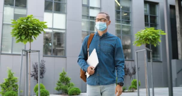 Retrato de un joven apuesto estudiante masculino en máscara médica y gafas de pie al aire libre que sostiene el ordenador portátil y mirando a la cámara. Concepto pandémico del Coronavirus. Caucásico hombre en la calle. — Vídeos de Stock