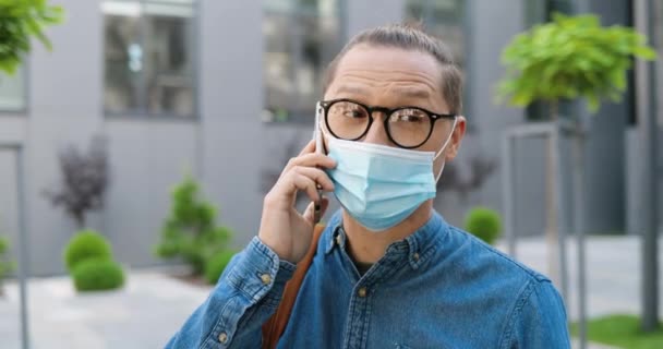 Dekat dengan pemuda Kaukasia berkacamata dan masker medis berbicara di ponsel di jalan kota. Pria tampan berkacamata berbicara lewat ponsel di luar ruangan. Percakapan panggilan telepon. — Stok Video