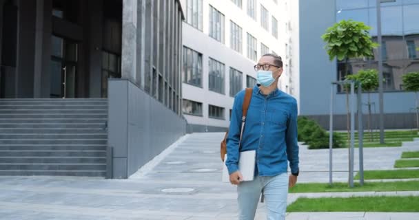 Portret młodego przystojnego studenta w masce medycznej i okularach spacerujących na świeżym powietrzu, trzymającego laptopa i patrzącego na boki. Koncepcja pandemii koronawirusowej. Kaukaski mężczyzna spacerujący po ulicy. — Wideo stockowe
